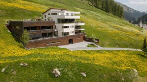 Ein modernes, mehrgeschossiges Gebäude mit Flachdach, umgeben von einer grünen Hügelslandschaft und einer Wiese mit gelben Blumen.
