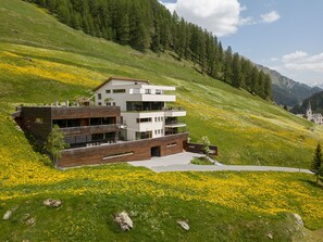Ein modernes, mehrgeschossiges Gebäude mit Flachdach, umgeben von einer grünen Hügelslandschaft und einer Wiese mit gelben Blumen.