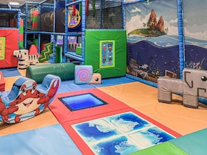 Ein Indoor-Spielbereich mit einer Spielkonstruktion, weichen Spielflächen und einer Wandmalerei mit Strandmotiv.