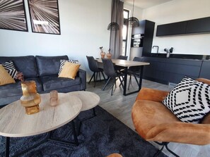 Ein modernes Wohnzimmer mit einer Couch, einem Couchtisch und einem Sessel, ein Essbereich mit Stühlen und eine Küche mit Schränken und einem Fernseher.