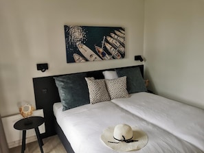 Ein ordentlich bezogenes Bett mit weißer Decke und einem Halmhut darauf. Darüber an der Wand hängt ein Gemälde mit Booten.