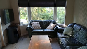 En stue med en sort lædersofa, et træbord, et fjernsyn monteret på væggen og et stort vindue med persienner.