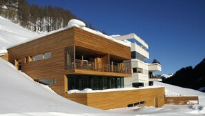 Un edificio moderno a più piani con pannelli in legno, balconi e ampie finestre, immerso in un pendio innevato.