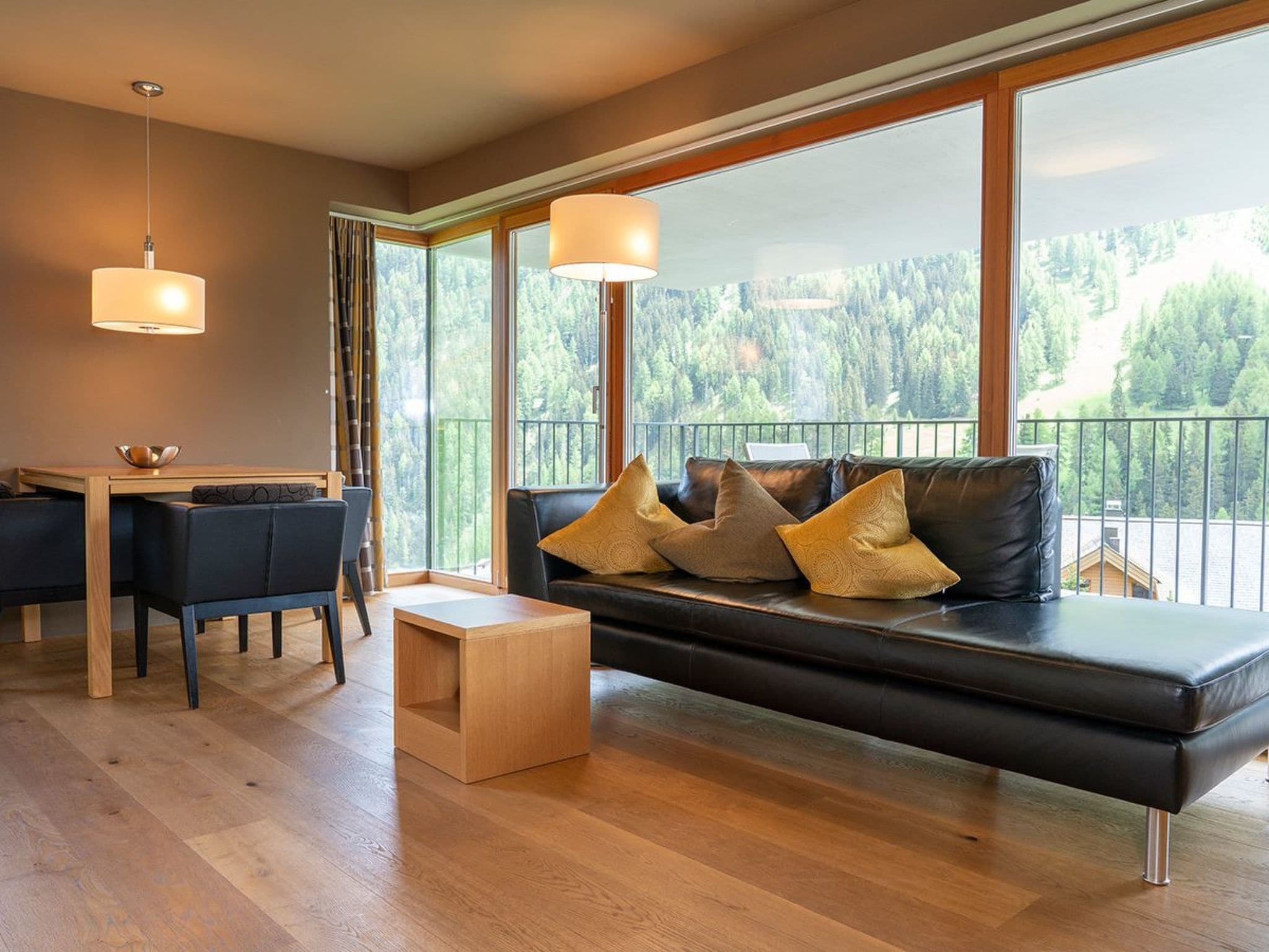 A modern living room with a sofa, a dining table, and a view of a forest through large windows.