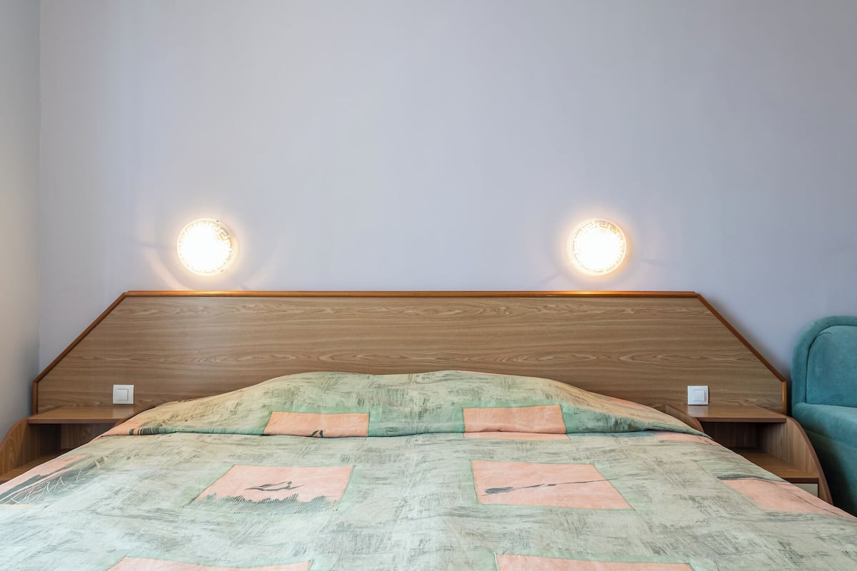 Ein Schlafzimmer mit einem Holz-Kopfteil, zwei Nachttischlampen, einem Bett mit gemustertem Überwurf und einem türkisfarbenen Sessel.