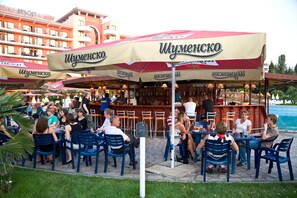 Zona de bar al aire libre con mesas y sillas, piscina al fondo y un edificio con balcones.