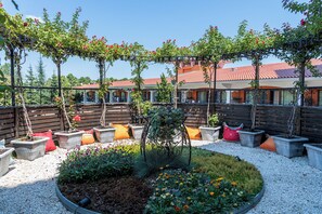 A courtyard with potted plants, a pergola with climbing plants, and a building with a red-tiled roof in the background. - Hrizantema (Nessebar)
