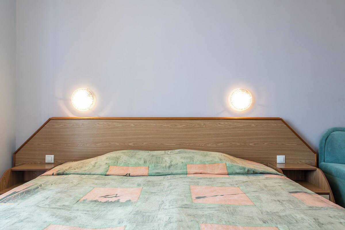 Un dormitorio con cabecera de madera, dos lámparas de noche, una cama con colcha estampada y una silla azul turquesa.