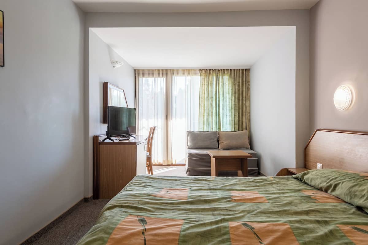 Ein Hotelzimmer mit einem Bett, einem Schreibtisch mit Computer, einem Fernseher, einer Couch und einem Fenster mit Vorhängen.