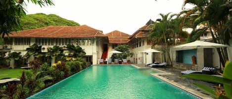 A swimming pool with lounge chairs, surrounded by palm trees and a building with a tiled roof.