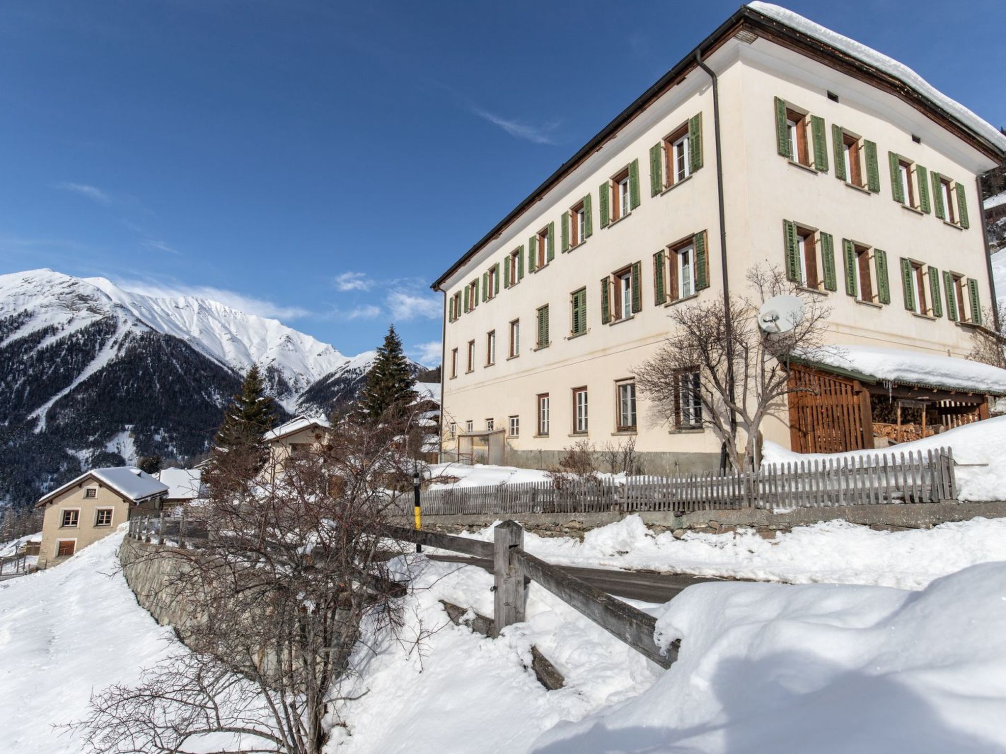Un edificio de varios pisos con tejado cubierto de nieve, rodeado de montañas y una valla de madera.