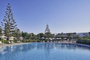 Una piscina con sillas de descanso y sombrillas, rodeada de árboles y edificios de hotel.