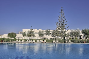 A pool with lounge chairs and umbrellas, surrounded by trees and a building with a modern design. - Atlantica Beach Resort (Kos)