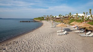 A beach with sun loungers and umbrellas, a clear sky, and buildings in the background. - Atlantica Beach Resort (Kos)