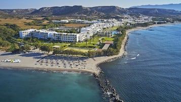 An aerial view of a coastal hotel with multiple buildings, a beach with sun loungers, and a rocky shoreline.