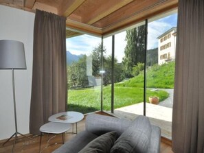 A room with a sofa, a small table, and a lamp. There is a large window with curtains, offering a view of a green landscape and a building. - Chasa Paoletta, (Ramosch) by Interhome (Ramosch)