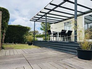 Ein überdachter Außenbereich mit Holzterrasse, einer Sitzgruppe und einem Gebäude mit Glasfassade.