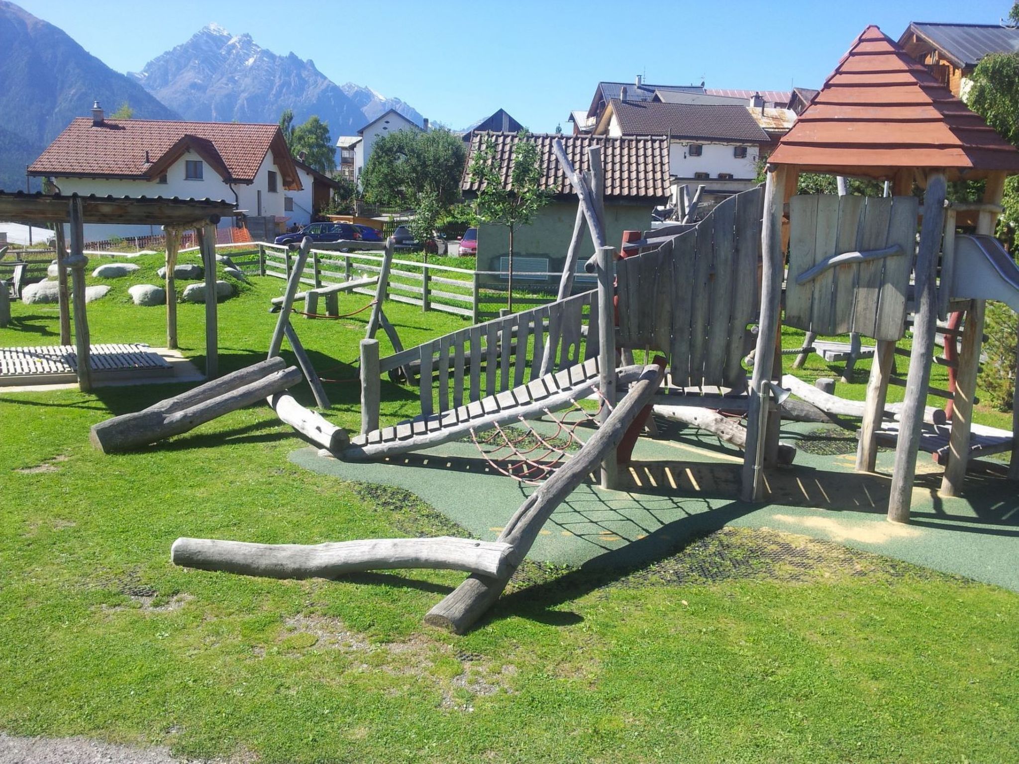 Un terrain de jeux avec des structures en bois et un espace vert, entouré de maisons et de montagnes.