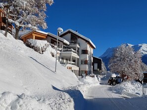 A snow-covered mountain road with buildings on the left, including a multi-story hotel with balconies and a ski lift visible in the background. - Ferienhof PUA - Erlebnisferien auf dem Bauernhof, (Sent) by Interhome (Sent)