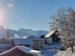 Un village enneigé, avec des bâtiments traditionnels, un clocher d’église et des montagnes en arrière-plan.
