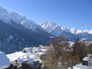 A mountainous landscape with snow-covered peaks and a small settlement with buildings. - Via Sura 577A, (Sent) by Interhome (Sent)