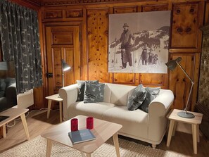 Ein gemütliches Wohnzimmer mit einer holzgetäfelten Wand, einem großen gerahmten Bild, einem weißen Sofa und einem Couchtisch mit Büchern und roten Tassen.