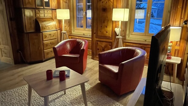 A cozy room with wooden paneling, a television, two leather chairs, a small table, and a window with a view of a snowy landscape.