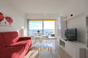 Un salón moderno con un sofá rojo, una mesa de centro de vidrio, un televisor y un balcón con vistas al mar.