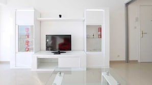 Ein modernes Wohnzimmer mit einem Flachbildfernseher auf einer weißen Unterhaltungseinheit, Glastrennwänden mit Gegenständen und einer Tür rechts.
