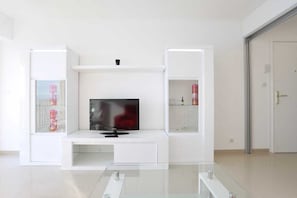 Un salon moderne avec un téléviseur à écran plat posé sur un meuble blanc, des étagères en verre ornées de divers objets, et une porte située à droite.