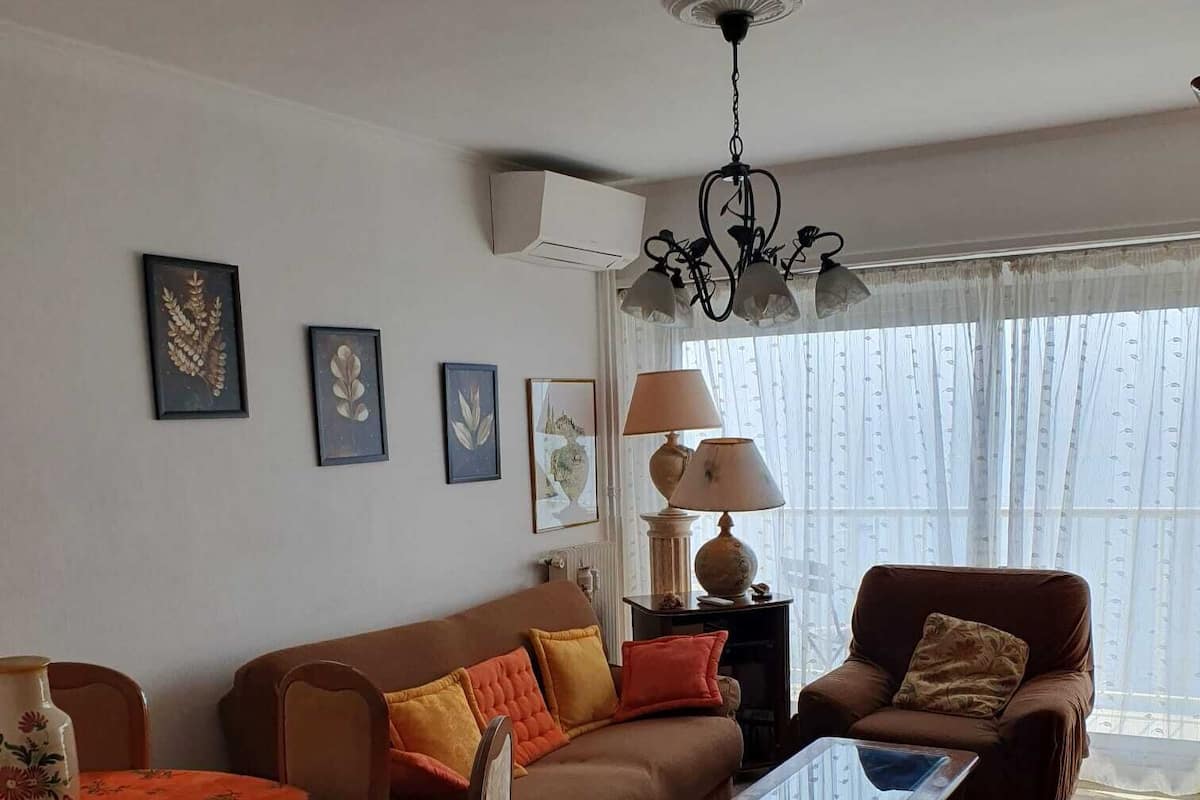Un salon avec un canapé marron, une table basse, une table à manger recouverte d’une nappe à motifs floraux et une lampe.