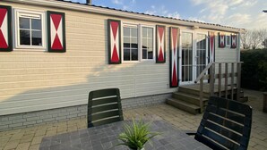 A mobile home with a brick foundation, a covered porch, and a small outdoor seating area with a table and chairs. - Rbr 813 - Beach Resort Kamperland by Interhome (Kamperland)