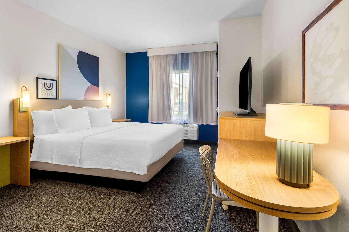 Ein Hotelzimmer mit Bett, Nachttisch, Lampe, Sessel, Fernseher und einem Bild an der Wand.