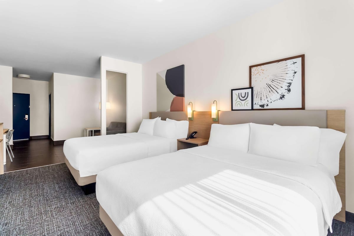 Ein Hotelzimmer mit zwei Betten, einem Schreibtisch, einem Stuhl, einem kleinen Tisch und einer Wand mit abstrakter Kunst.