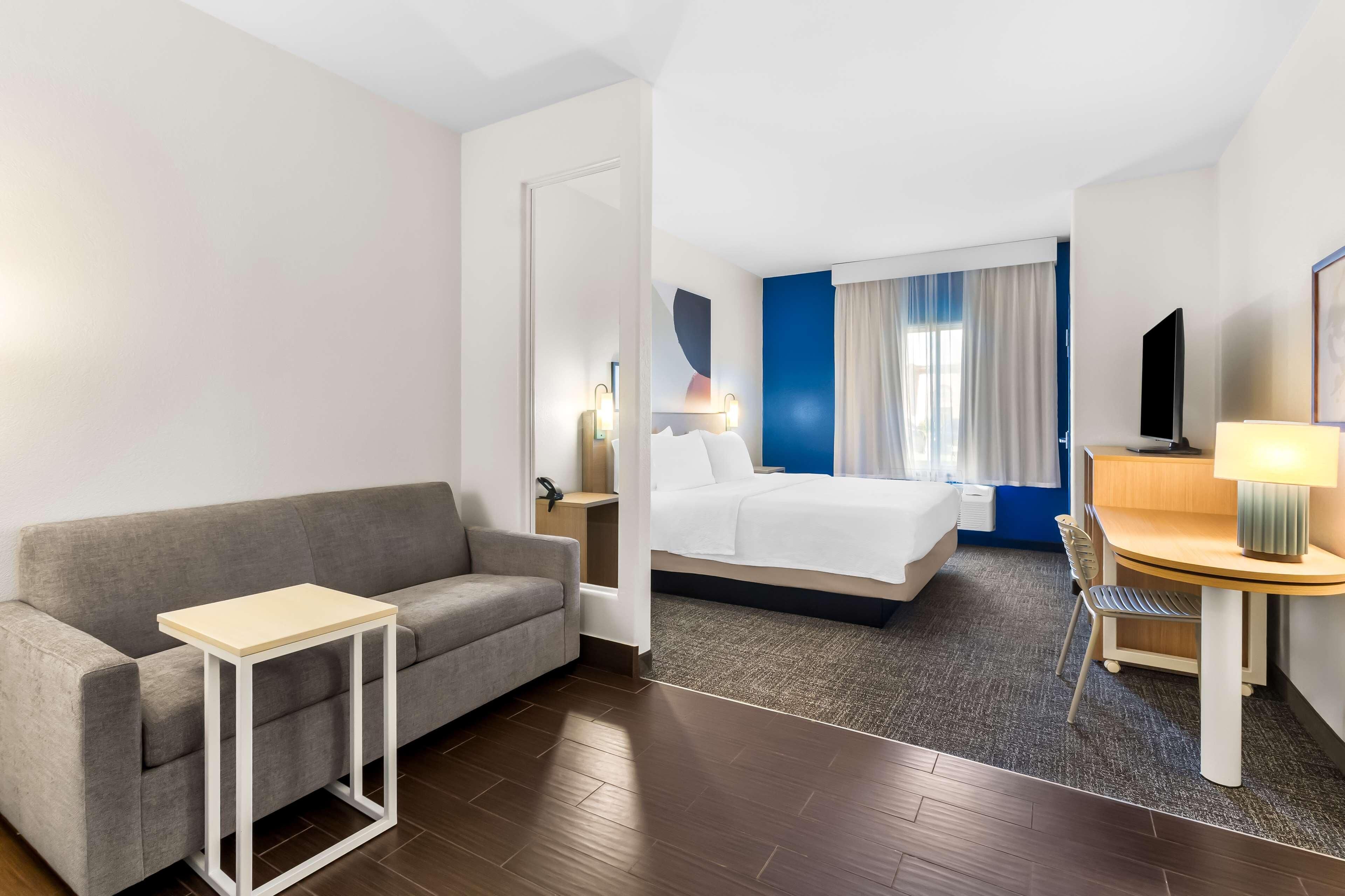 Ein Hotelzimmer mit einer Couch, einem kleinen Tisch, einem Bett, einem Schreibtisch, einem Sessel, einem Fernseher und einem Fenster mit Vorhängen.