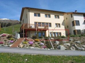 A two-story building with a balcony, a garden with colorful flowers, and a hilly background.