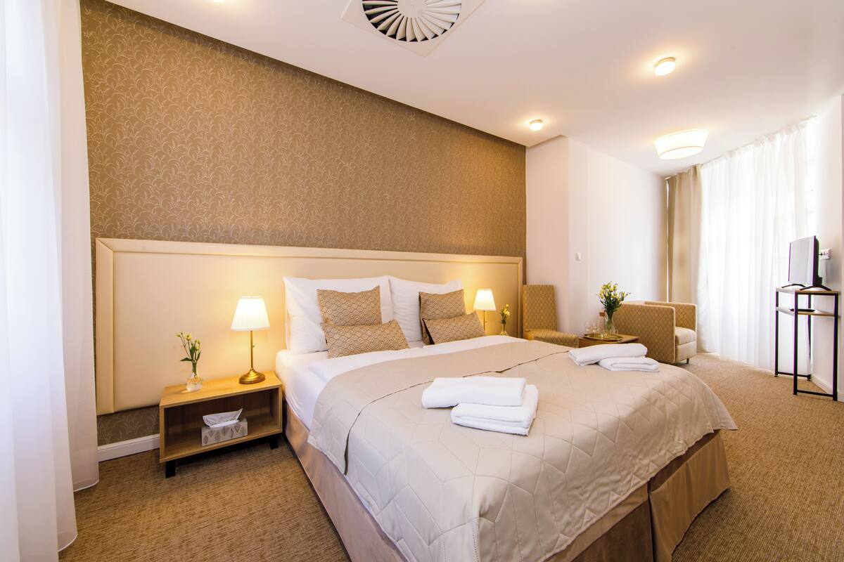 Una habitación de hotel con una cama grande, mesitas de noche, una silla, un televisor y una ventana con cortinas.