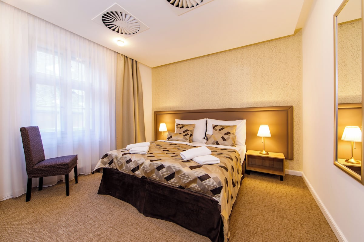 Una habitación de hotel con cama, una silla, dos mesitas de noche con lámparas y un ventanal con cortinas.