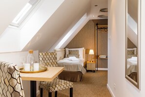 Habitación de hotel con techo inclinado, una cama, una mesita de noche con lámpara, una pequeña mesa con vasos y una botella, y un espejo en la pared.