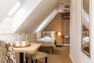 Quarto de hotel com teto inclinado, uma cama, mesa de cabeceira com abajur, pequena mesa com copos e uma garrafa, e um espelho na parede.