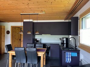 Una cucina compatta con mobili scuri, una zona pranzo con un tavolo di legno e sedie nere, e illuminazione a sospensione.