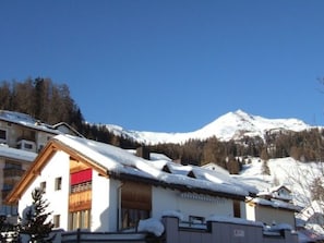 A snow-covered mountain with a clear sky, a building with a red awning, and a wooden door. - Chasa Cumulus/HP Ferienwohnung Sainas 139 A, (Ftan) by Interhome (Ftan)
