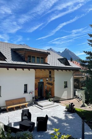 Ein zweigeschossiges Gebäude mit Holzfassade, einer überdachten Veranda und Gartenmöbeln, vor einer Bergkulisse.