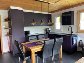Eine moderne Küche mit dunklen Schränken, einer zentralen Kochinsel, Pendelleuchten und einem Essbereich mit Holztisch und Stühlen.