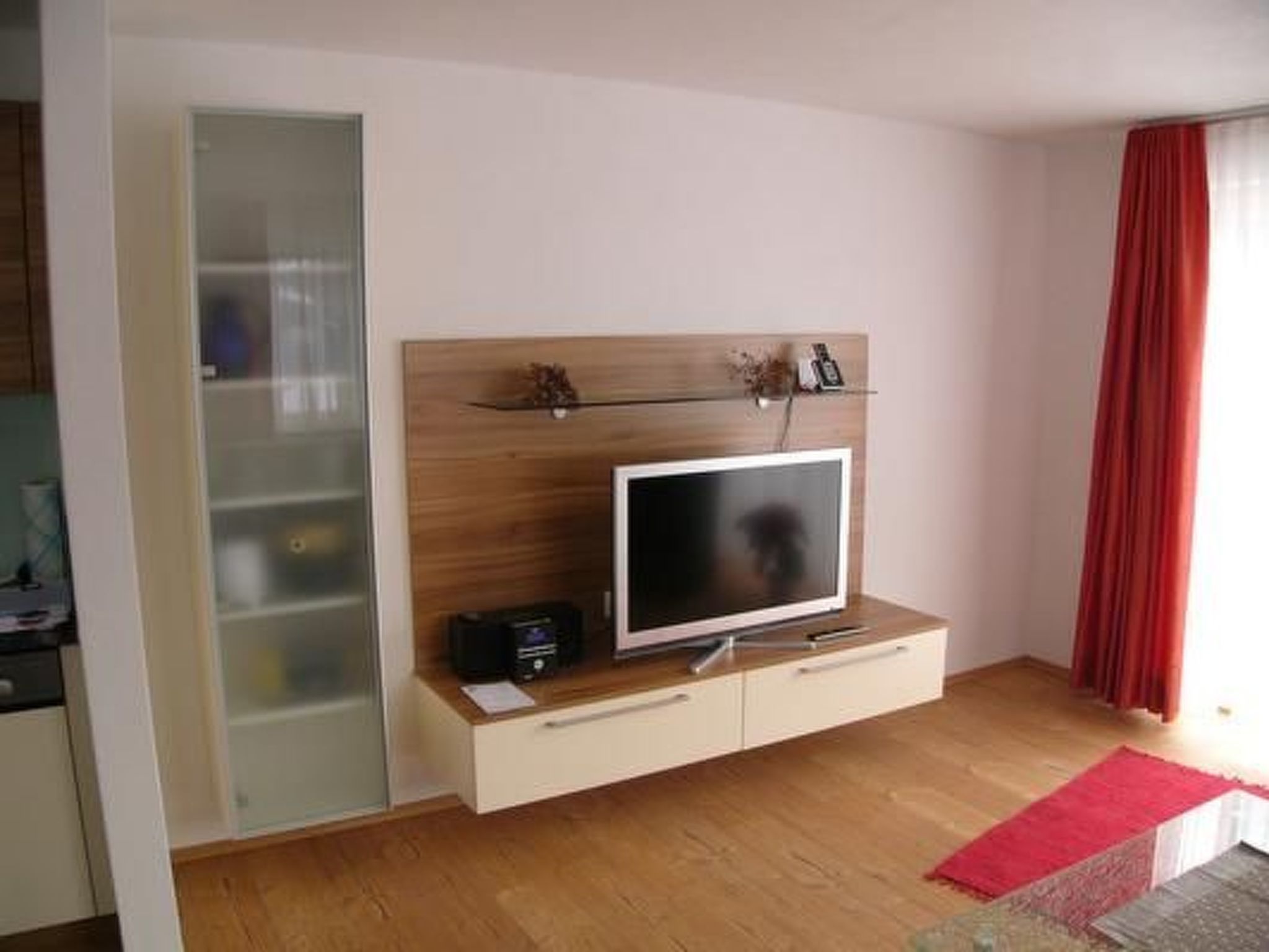 Una sala moderna con un televisor de pantalla plana sobre un mueble de madera, un aparador con puertas de vidrio a la izquierda y una cortina roja a la derecha.