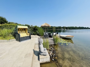 Eine friedliche Uferlandschaft mit einem Gazebo mit Reetdach, einer steinernen Buddha-Statue und einem kleinen Boot, das am Wasser liegt.