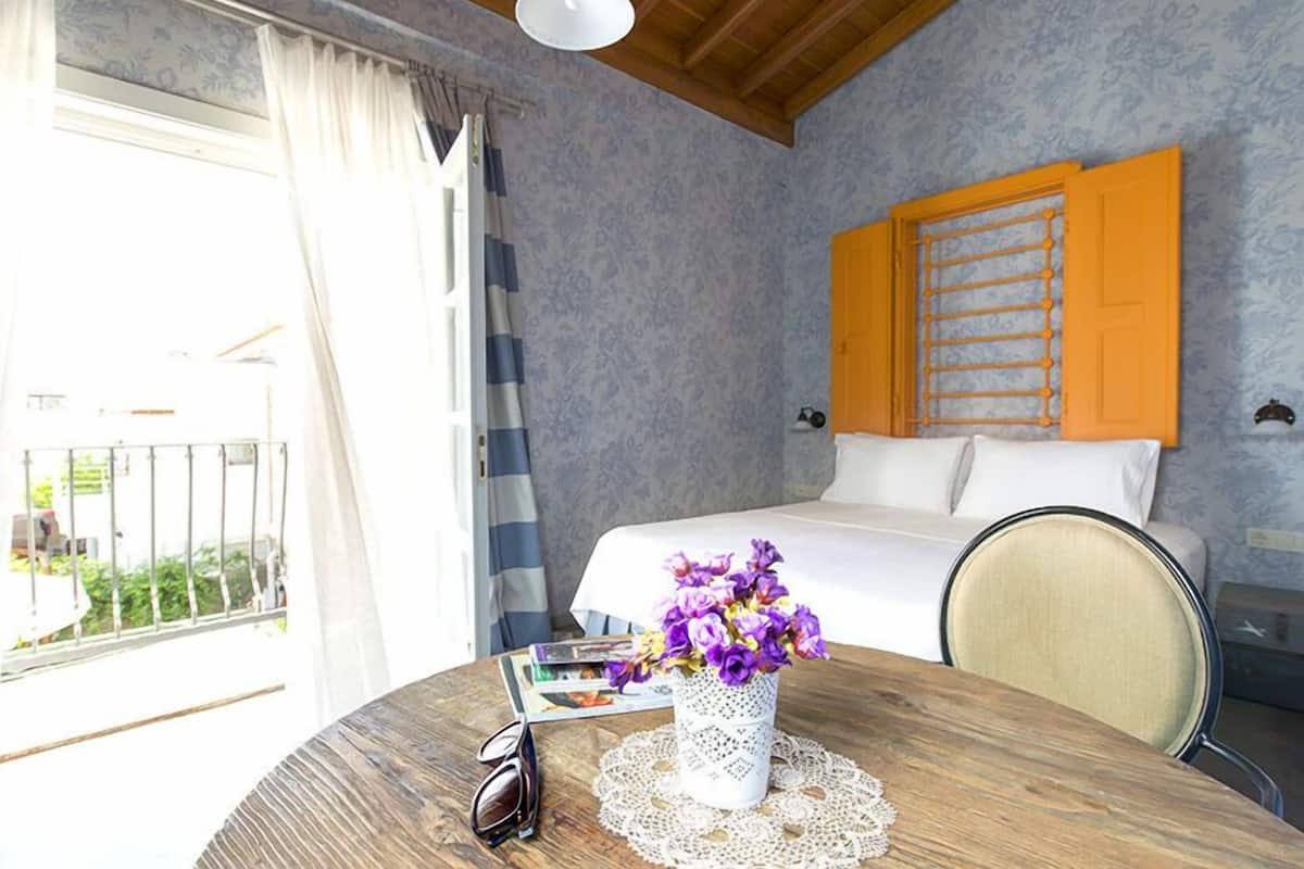 A hotel room with a bed, a table with a vase of flowers, and a view of the outside.