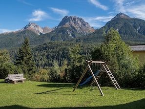 A grassy area with a wooden swing set and a bench, set against a backdrop of mountains and trees. - WÜEST, (Scuol) by Interhome (Scuol)