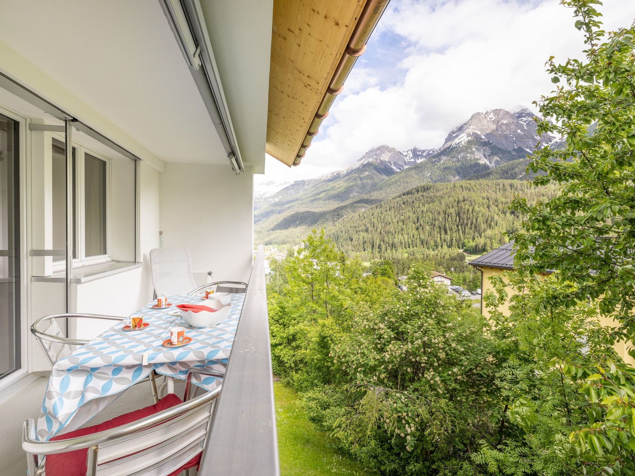 Un balcon avec une table dressée pour le dîner, offrant une vue sur un paysage montagneux, avec des arbres et un village au loin.