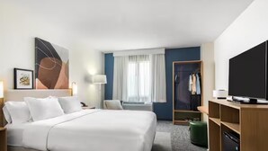Habitación de hotel con cama, escritorio, televisor, armario y ventana con cortinas.